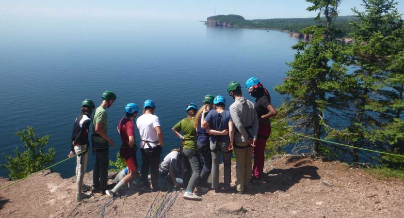 A group of students wearing safety gear stand on a rocky overlook above a vast blue lake. 